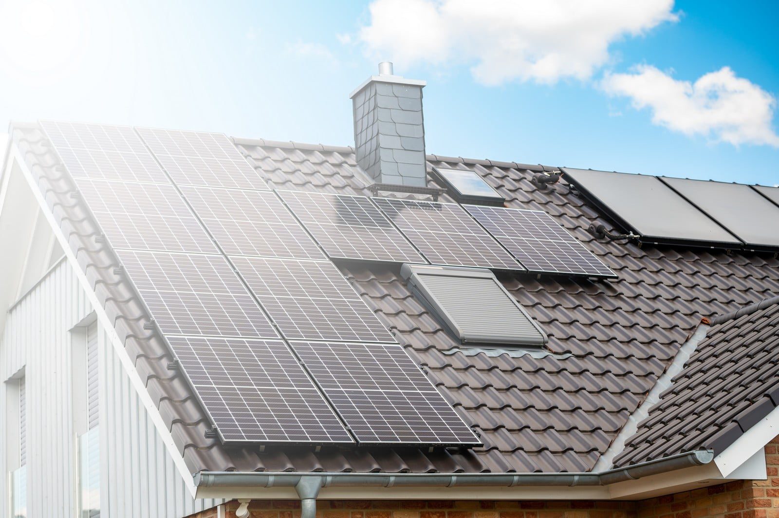 The image depicts a modern residential house with a roof fully equipped with an array of solar panels, capturing sunlight to generate renewable energy. The panels are symmetrically arranged, covering the majority of the roof’s surface. The house itself appears to be constructed with a combination of sleek, contemporary materials, featuring a predominantly light-coloured exterior with large, energy-efficient windows reflecting the clear blue sky. The solar panels have a dark hue, contrasting sharply with the lighter rooftop beneath them. This setup exemplifies an eco-friendly approach to harnessing solar energy, with the house likely positioned to maximize sun exposure. The surrounding area hints at a suburban setting, with trees and neighboring houses faintly visible in the background. The overall scene exudes a sense of environmental stewardship and modern sustainable living.