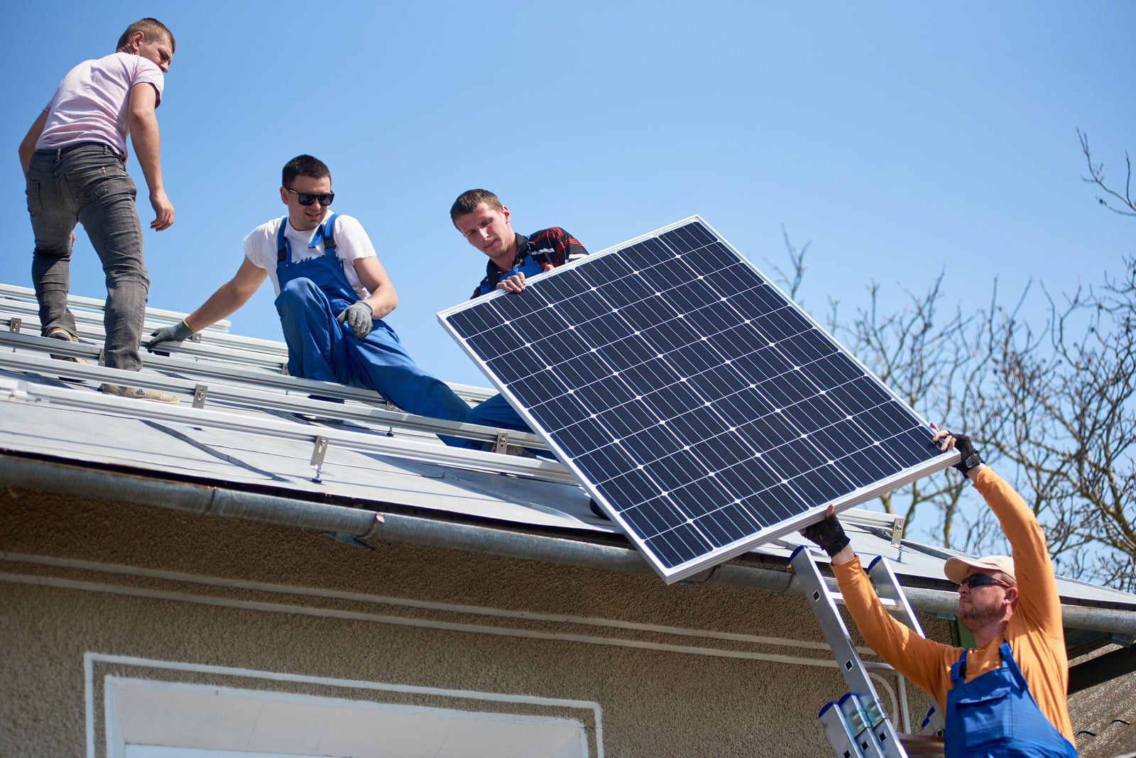 The image shows a team of workers installing solar photovoltaic panels on a sloped rooftop on a bright, sunny day. The roof is covered in solar panel arrays, which are being methodically arranged in neat rows. The workers, wearing safety helmets and reflective vests for safety, are actively engaged in positioning and securing the solar panels. One worker is kneeling with a tool in hand, appearing to tighten a fixture, while another stands nearby, holding a panel ready for installation. The background includes a clear blue sky and a distant treeline, indicating a suburban or rural area. The scene highlights both the teamwork and the technical expertise involved in setting up a solar power system, emphasising the shift towards renewable energy sources.