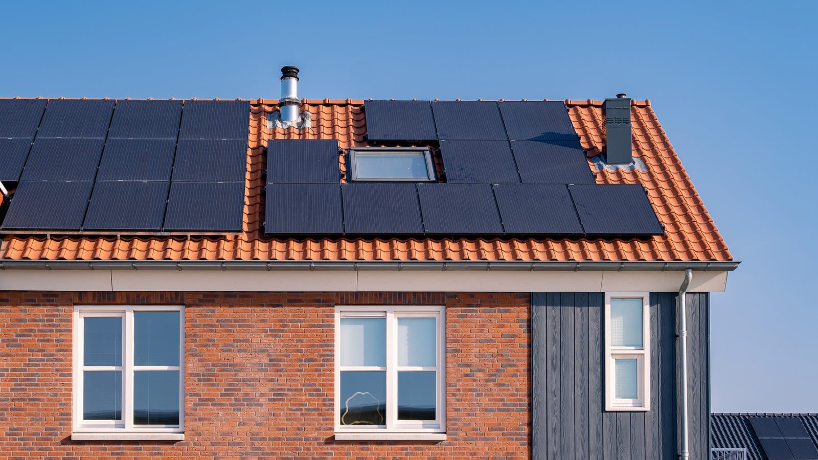 The image depicts a modern residential neighborhood comprising newly built houses. Each house is constructed with a contemporary architectural design, featuring clean lines, large windows, and a blend of neutral tones such as white, grey, and beige. The roofs of these houses are equipped with solar panels, indicating a commitment to sustainable energy use. The solar panels are neatly aligned and cover a substantial portion of each roof, maximising exposure to sunlight. The surroundings are well-maintained, with neatly trimmed green lawns, young trees, and shrubbery lining the streets, which are wide and smoothly paved. The overall ambiance of the scene is serene, under a clear blue sky with a few scattered clouds, suggesting a bright, sunny day. This neighborhood reflects modern living with an emphasis on eco-friendliness and sustainability.