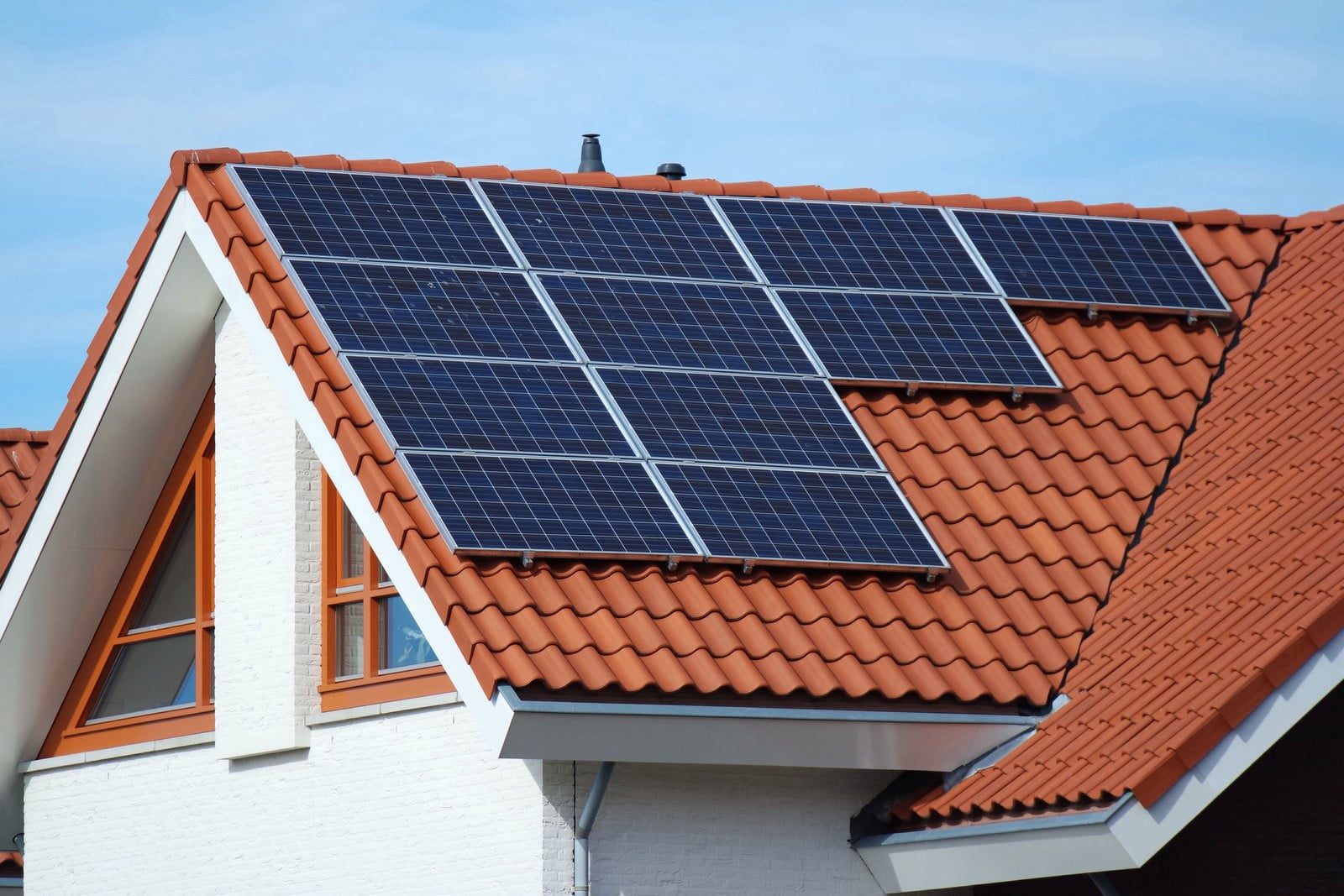 The image depicts a rooftop array of modern solar panels installed on a residential building under a clear, blue sky. Each rectangular panel is neatly aligned, reflecting sunlight, indicating efficient positioning for maximum solar energy absorption. The solar panels are a blend of dark blue and black hues, typical of photovoltaic cells, which are framed in metallic borders to enhance durability and stability. The rooftop is constructed from a dark, textured material, possibly asphalt shingles, providing a sturdy foundation for the solar panels. In the background, a few distant clouds drift gently across the sky, emphasising the serene and unobstructed environment ideal for solar energy harvesting. The overall ambiance of the image highlights clean, renewable energy solutions, suggesting a commitment to sustainability and eco-friendly practices.